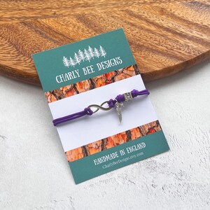 May include: A purple cord bracelet with an infinity symbol and a feather charm. The bracelet is displayed on a card with the text "CHARLY BEE DESIGNS" and "MADE IN ENGLAND". The background is a wooden surface.