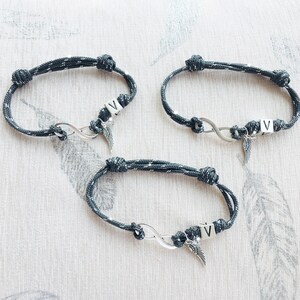 K&ouml;nnte beinhalten: Drei graue und silberne Seilarmb&auml;nder mit Unendlichkeitssymbolen und dem Buchstaben "V". Jedes Armband hat einen kleinen silbernen Fl&uuml;gelanh&auml;nger. Die Armb&auml;nder sind geknotet und scheinen handgefertigt zu sein.