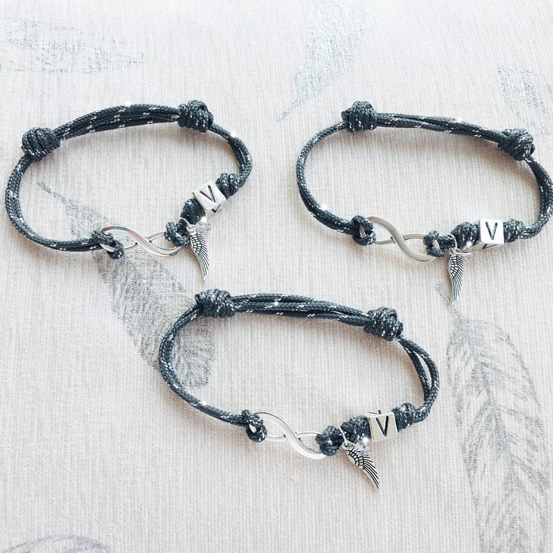 K&ouml;nnte beinhalten: Drei graue und silberne Seilarmb&auml;nder mit Unendlichkeitssymbolen und dem Buchstaben "V". Jedes Armband hat einen kleinen silbernen Fl&uuml;gelanh&auml;nger. Die Armb&auml;nder sind geknotet und scheinen handgefertigt zu sein.