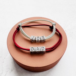 Puede incluir: Dos pulseras personalizadas con cuentas cúbicas plateadas que deletrean "JOE" y "DAD". Una pulsera tiene un cordón rojo y blanco, la otra un cordón rojo. Las pulseras se exhiben sobre una superficie redonda rosa.