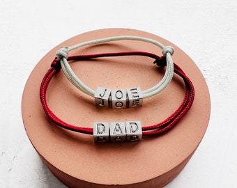 Puede incluir: Dos pulseras personalizadas con cuentas cúbicas plateadas que deletrean "JOE" y "DAD". Una pulsera tiene un cordón rojo y blanco, la otra un cordón rojo. Las pulseras se exhiben sobre una superficie redonda rosa.