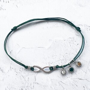 Op de afbeelding: Enkelband met een donkergroen koord, een zilveren oneindigheidssymbool en drie bungelende bedels. De bedels zijn een helder kristal, een groen kristal en een goudkleurig kristal. De armband is verstelbaar.