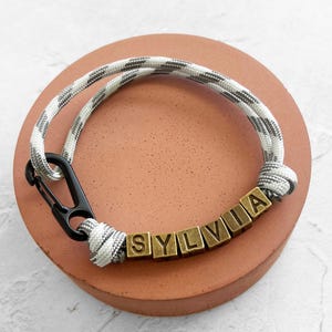 May include: A personalized bracelet with the name "SYLVIA" spelled out in gold-colored letter beads. The bracelet features a gray and white patterned rope and a black clasp. The bracelet is displayed on a round, reddish-brown surface.