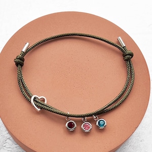 Puede incluir: Una pulsera de cordón verde oliva con un dije de corazón plateado y tres colgantes de piedras preciosas en rojo, rosa y azul. La pulsera está anudada en los extremos con detalles metálicos plateados.