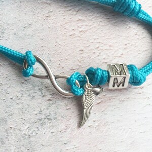 K&ouml;nnte beinhalten: T&uuml;rkisfarbenes Seilarmband mit einem silbernen Unendlichkeitssymbol, einem kleinen Fl&uuml;gelanh&auml;nger und einem W&uuml;rfel mit den Buchstaben "MM". Das Armband ist geknotet und hat ein nautisches Thema.