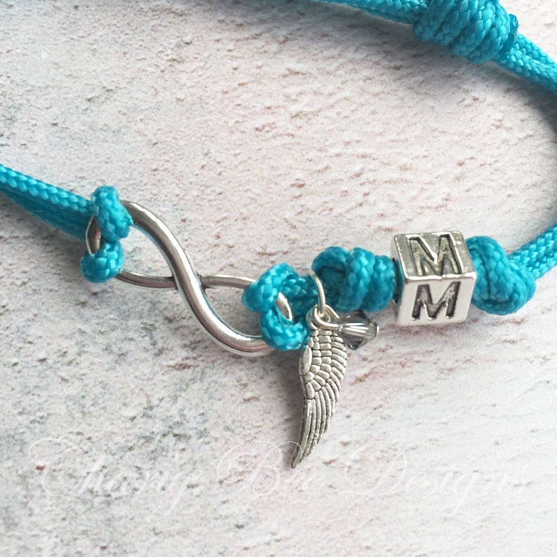 K&ouml;nnte beinhalten: T&uuml;rkisfarbenes Seilarmband mit einem silbernen Unendlichkeitssymbol, einem kleinen Fl&uuml;gelanh&auml;nger und einem W&uuml;rfel mit den Buchstaben "MM". Das Armband ist geknotet und hat ein nautisches Thema.
