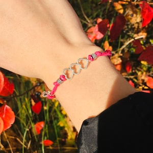 May include: A pink bracelet with three silver heart charms. The bracelet is made of a woven pink cord with silver beads on each side of the hearts. The background shows red leaves and green foliage.