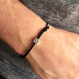 Puede incluir: Pulsera de cord&oacute;n negro con una cuenta cuadrada plateada con un dise&ntilde;o circular. La pulsera est&aacute; anudada para ajustarla y tiene extremos met&aacute;licos plateados. Un accesorio minimalista.
