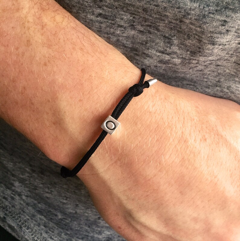 Puede incluir: Pulsera de cord&oacute;n negro con una cuenta cuadrada plateada con un dise&ntilde;o circular. La pulsera est&aacute; anudada para ajustarla y tiene extremos met&aacute;licos plateados. Un accesorio minimalista.