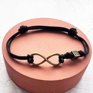 May include: Black cord bracelet featuring a bronze-colored infinity symbol and a small cube with the letter "P". The bracelet is knotted for adjustability. A simple, elegant piece of jewelry.