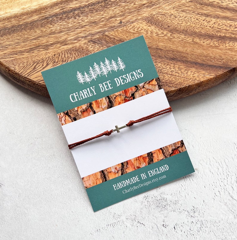 May include: A brown cord bracelet with a small silver cross charm, displayed on a teal card with the text "Charly Bee Designs" and "Made in England". The card has a wood grain pattern.