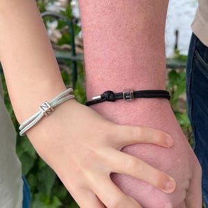 Puede incluir: Dos pulseras a juego. Una es azul claro con un dije cuadrado plateado con la letra "N". La otra es negra con un dije cuadrado plateado con la letra "E". Las pulseras se llevan en las muñecas.