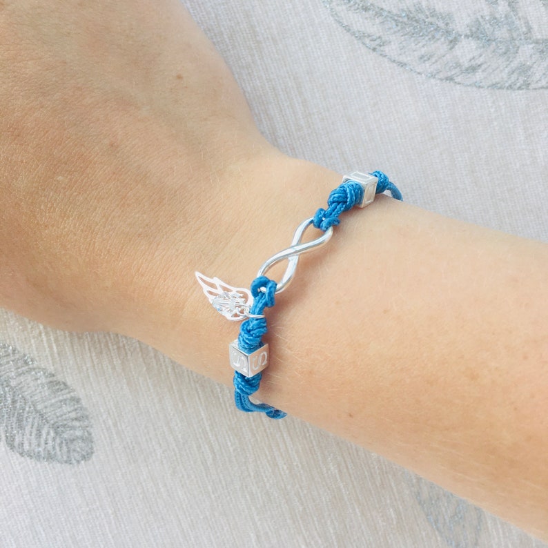 May include: A blue cord bracelet with silver-toned charms. The bracelet features an infinity symbol, a pair of wings, and two small square beads. The bracelet is worn on a wrist.