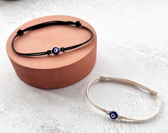 May include: Two adjustable cord bracelets with evil eye charms. One bracelet is black, and the other is off-white. The evil eye charms have a blue iris, white highlights, and a black pupil. The bracelets are displayed on a pink block.