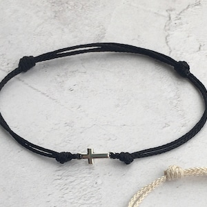May include: Black cord bracelet featuring a small silver cross charm. The bracelet is adjustable with sliding knots. The design is simple and minimalist, suitable for everyday wear. The bracelet is shown against a neutral background.