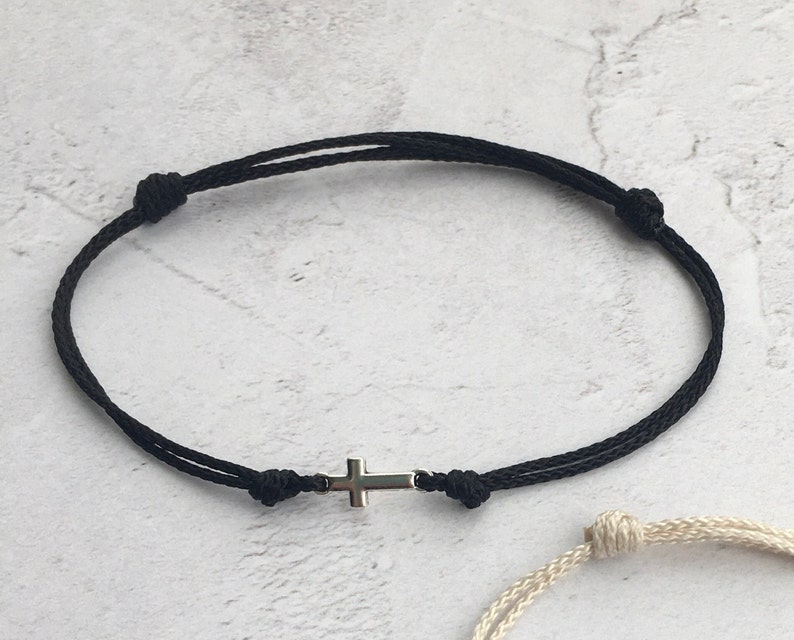 May include: Black cord bracelet featuring a small silver cross charm. The bracelet is adjustable with sliding knots. The design is simple and minimalist, suitable for everyday wear. The bracelet is shown against a neutral background.