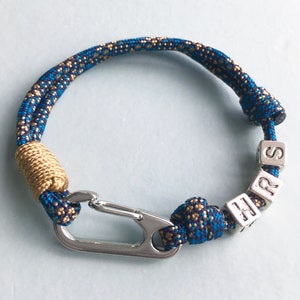 Könnte beinhalten: Ein blau-goldenes Seilarmband mit einem silbernen Verschluss und silbernen Buchstabenwürfeln mit der Aufschrift "SHRS". Das Armband hat einen goldfarbenen Abschnitt in der Nähe des Verschlusses.