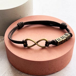 May include: A brown cord bracelet featuring an infinity symbol and three lettered cubes. The infinity symbol and cubes are in a bronze color. The bracelet is displayed on a pink surface.
