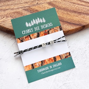 Puede incluir: Una pulsera de cord&oacute;n blanco y negro con cuentas de letras plateadas que deletrean "AB" se muestra en una tarjeta verde y blanca. La tarjeta presenta el texto "Charly Bee Designs" y "Handmade in England."