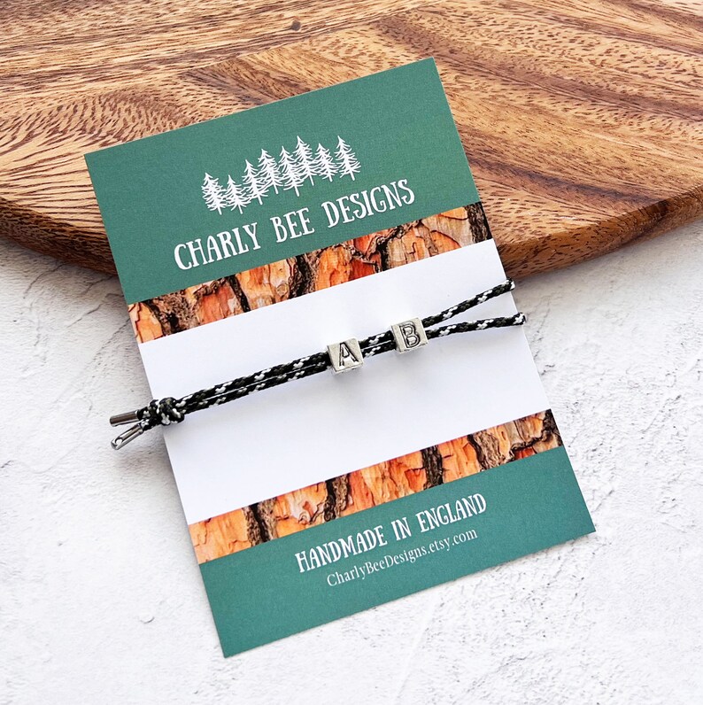 Puede incluir: Una pulsera de cord&oacute;n blanco y negro con cuentas de letras plateadas que deletrean "AB" se muestra en una tarjeta verde y blanca. La tarjeta presenta el texto "Charly Bee Designs" y "Handmade in England."