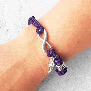 May include: A purple infinity bracelet with a silver charm. The bracelet features a braided cord design and a silver infinity symbol. A small silver charm with wings and a square silver bead are attached.