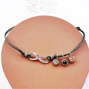 May include: A silver-toned infinity symbol bracelet with a dark green cord. The bracelet features three small, round charms with clear, green, and yellow gemstones. The infinity symbol is the central design element.