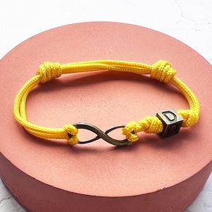 May include: Yellow cord bracelet with an infinity symbol and a cube bearing the letter "D". The bracelet is knotted at each end. The infinity symbol is bronze-coloured. The bracelet rests on a pink surface.