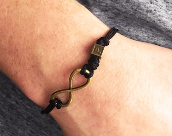Puede incluir: Pulsera de cordón negro con un símbolo de infinito de color bronce y un pequeño acento cuadrado. La pulsera está anudada y es ajustable. El símbolo del infinito es un diseño clásico que simboliza la eternidad.