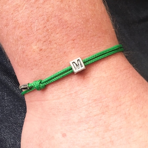 Puede incluir: Una pulsera de cord&oacute;n verde con una cuenta cuadrada plateada con la letra "M". La pulsera est&aacute; anudada y tiene un cierre de metal. La pulsera se lleva en la mu&ntilde;eca.