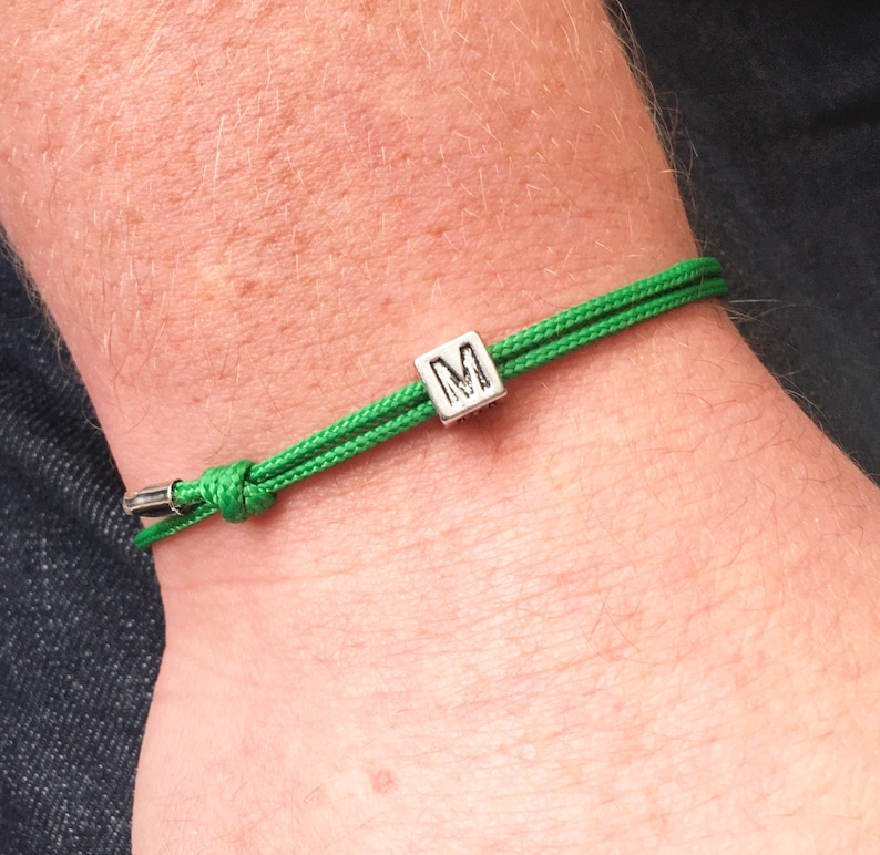 Puede incluir: Una pulsera de cord&oacute;n verde con una cuenta cuadrada plateada con la letra "M". La pulsera est&aacute; anudada y tiene un cierre de metal. La pulsera se lleva en la mu&ntilde;eca.