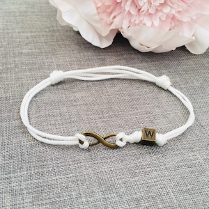 Può includere: Braccialetto con cordino bianco con un simbolo infinito color bronzo e una piccola perla quadrata con la lettera "W". Il braccialetto è annodato e regolabile. Un fiore rosa chiaro è sullo sfondo.