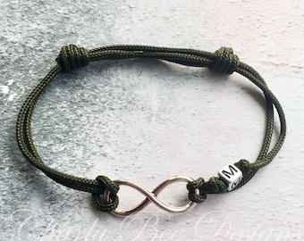 Puede incluir: Pulsera ajustable verde oliva con un símbolo de infinito plateado y una pequeña cuenta cúbica con la letra "M". La pulsera está hecha de un cordón tejido y es ajustable. El símbolo del infinito es un diseño clásico.