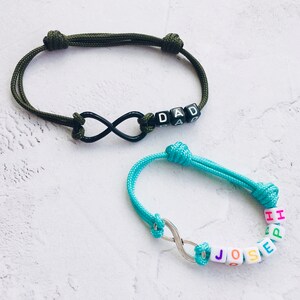 Puede incluir: Dos pulseras de infinito hechas a mano. Una es verde oliva con cuentas negras que deletrean "DAD". La otra es turquesa con cuentas que deletrean "JOSE" y "HARPER". Ambas pulseras presentan un símbolo de infinito.