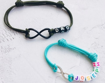 Puede incluir: Dos pulseras de infinito hechas a mano. Una es verde oliva con cuentas negras que deletrean "DAD". La otra es turquesa con cuentas que deletrean "JOSE" y "HARPER". Ambas pulseras presentan un símbolo de infinito.