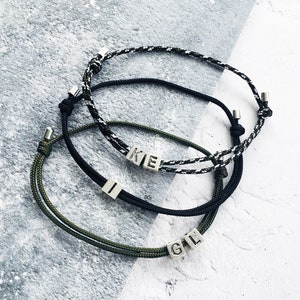 Op de afbeelding: Drie verstelbare armbanden met gepersonaliseerde letterkralen. Eén armband is zwart-wit, één is zwart en één is olijfgroen. De armbanden liggen op een lichtgrijs en wit oppervlak.