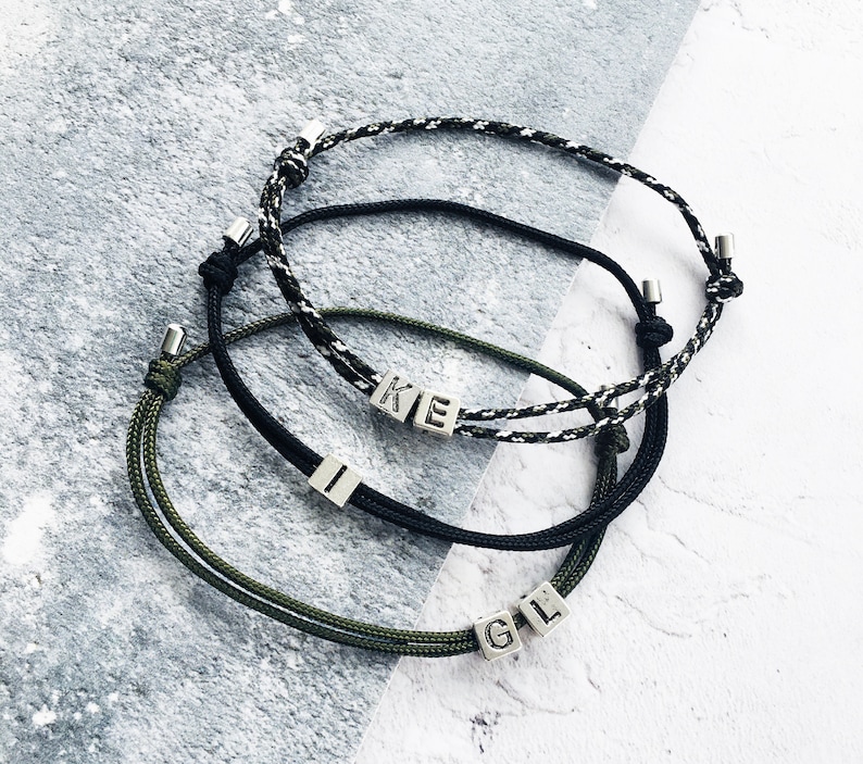 Puede incluir: Tres pulseras ajustables con cuentas de letras personalizadas. Una pulsera es en blanco y negro, otra en negro y la otra en verde oliva. Las pulseras est&aacute;n sobre una superficie gris y blanca.