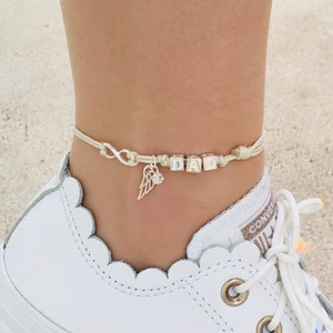 Könnte beinhalten: Cremefarbenes Fußkettchen mit silbernen Anhängern. Das Armband buchstabiert "DAD" mit silbernen Buchstabenperlen. Es hat auch ein Unendlichkeitssymbol und einen Flügel-Charm.