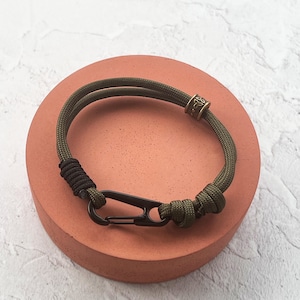 May include: Olive green paracord bracelet with a black clasp and accents. The bracelet features a decorative bronze-colored bead and a black wrapped section. The bracelet is displayed on a round, reddish-orange surface.