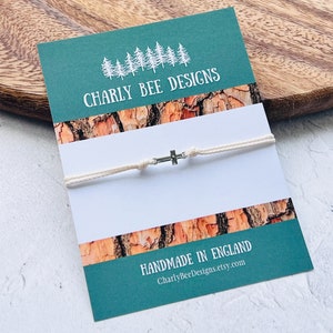 May include: A cream-colored cord bracelet with a small silver cross charm, displayed on a teal card with the text "CHARLY BEE DESIGNS" and "HANDMADE IN ENGLAND". The card has a wood-grain pattern.