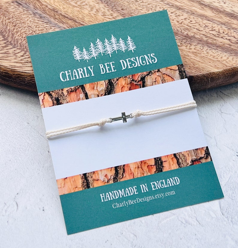 May include: A cream-colored cord bracelet with a small silver cross charm, displayed on a teal card with the text "CHARLY BEE DESIGNS" and "HANDMADE IN ENGLAND". The card has a wood-grain pattern.