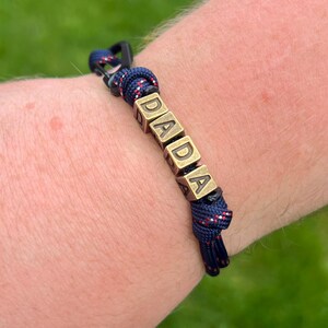 May include: A navy blue bracelet with the word "DADA" spelled out in gold-colored square beads. The bracelet is made of a woven rope with red and white accents. The clasp is black.