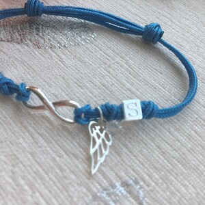 May include: A blue cord bracelet featuring a silver infinity symbol, a small wing charm, and a white cube with the letter "S". The bracelet is knotted and adjustable.