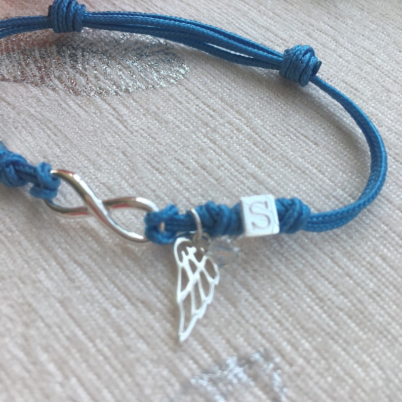 May include: A blue cord bracelet featuring a silver infinity symbol, a small wing charm, and a white cube with the letter "S". The bracelet is knotted and adjustable.