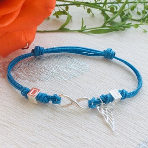 May include: A turquoise adjustable bracelet featuring a silver infinity symbol and a small wing charm. The bracelet has two silver square beads with the letters "D" and "S". The bracelet is tied with knots on each end.