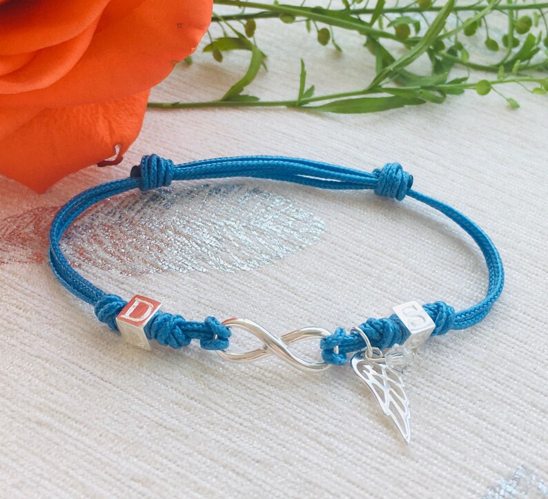 May include: A turquoise adjustable bracelet featuring a silver infinity symbol and a small wing charm. The bracelet has two silver square beads with the letters "D" and "S". The bracelet is tied with knots on each end.