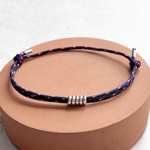 Puede incluir: Pulsera de estilo náutico con un diseño de cuerda azul marino, roja y blanca. La pulsera tiene una serie de anillos de metal plateado en el centro y extremos de metal plateado. Se muestra sobre una superficie de color terracota.