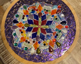 May include: A round wooden tray with a purple mosaic design. The mosaic features a variety of colorful glass and ceramic pieces, including red, orange, yellow, green, blue, and purple. The center of the mosaic features a starburst design.