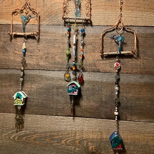 May include: Three handcrafted wind chimes with copper wire frames and beaded accents. Each features a bird or birdhouse design, with colorful beads and charms dangling below. The background is a rustic wooden wall.