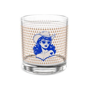 May include: A clear glass tumbler featuring a blue graphic of a person wearing a cowboy hat and heart-shaped sunglasses. The background pattern consists of small, repeating tan-coloured icons of a hand making a middle finger gesture.