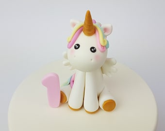 May include: A white fondant unicorn cake topper with a gold horn, rainbow mane, and pink wings. The unicorn is sitting on a white cake with a pink number one.